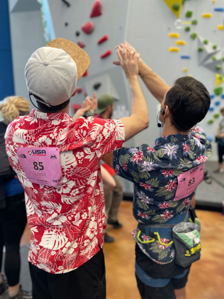 Matt Fredrick (left) stands beside Justin Salas holding Salas’ wrists, guiding his arms and hands through the movement sequence of the climb in front of them at the 2023 Paraclimbing National Championship in Austin, Texas. (Image credit: Emily Chen-Newton)