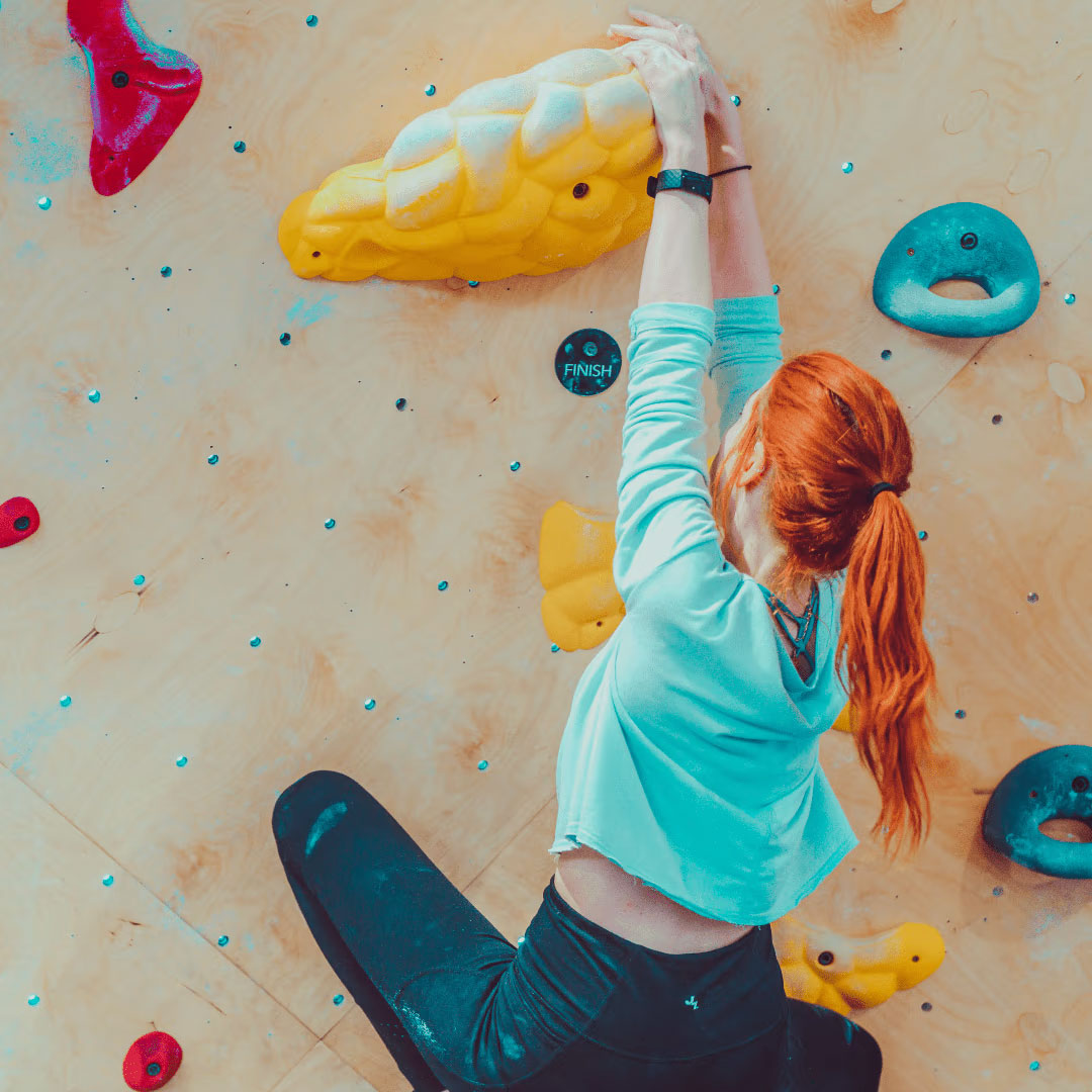 A climber with a turquoise hoodie and bright red ponytail reaches high to grab a lumpy yellow hold resembling a loaf french brioche bread. The yellow holds below her share this shape and texture as a prime example of setting similar holds together. (Image credit: Oso Climbing Gym)